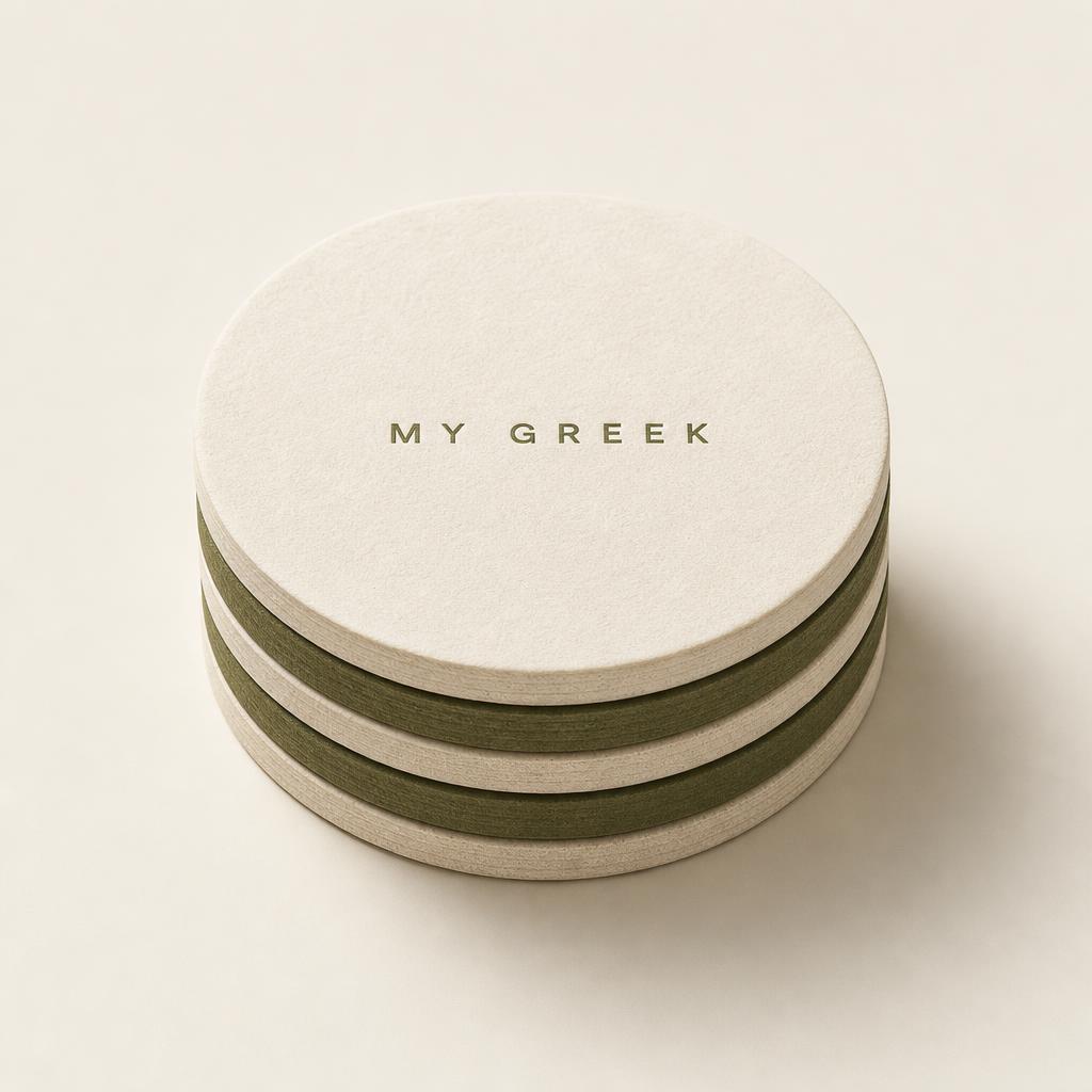 Stack of cream and olive letterpress paper coasters printed with the MY GREEK mark